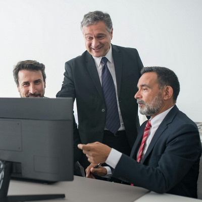 pexels-kampus-8353834-400 Three men looking at a computer screen, two of them sitting, one standing up between the other two. The man on the right is pointing at the screen. We cannot see the screen.