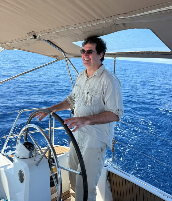 2025-06-01 Keith navigating a boat 560x653 Keith Burman at the helm of a sailboat. He is wearing sunglasses.